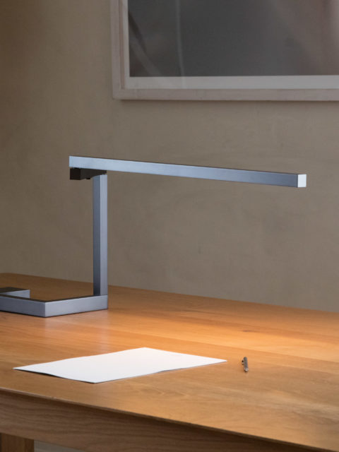 Untitled S gunmetal gray table lamp illuminates a sheet of paper on a wooden desk