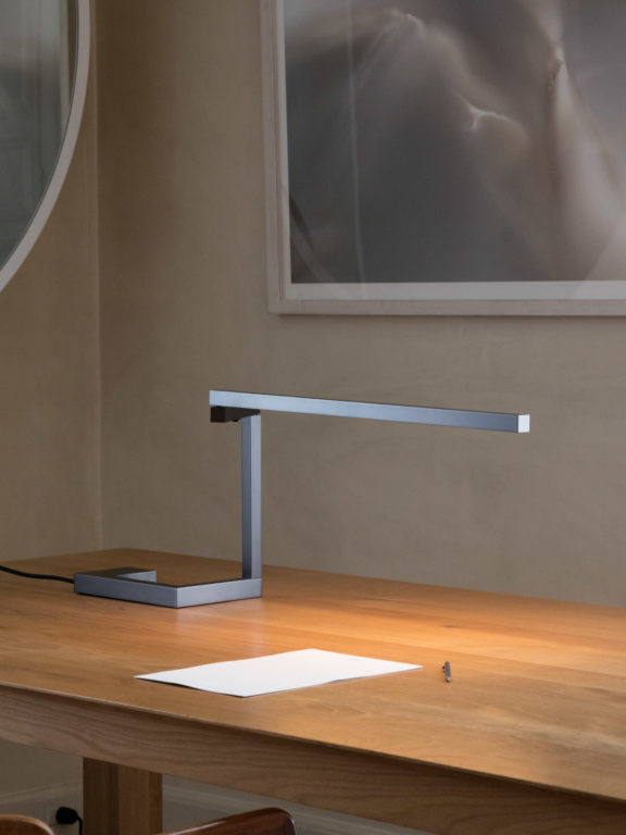 Die Tischlampe Untitled S in der Farbe Gunmetal beleuchtet ein Blatt Papier auf einem Holztisch