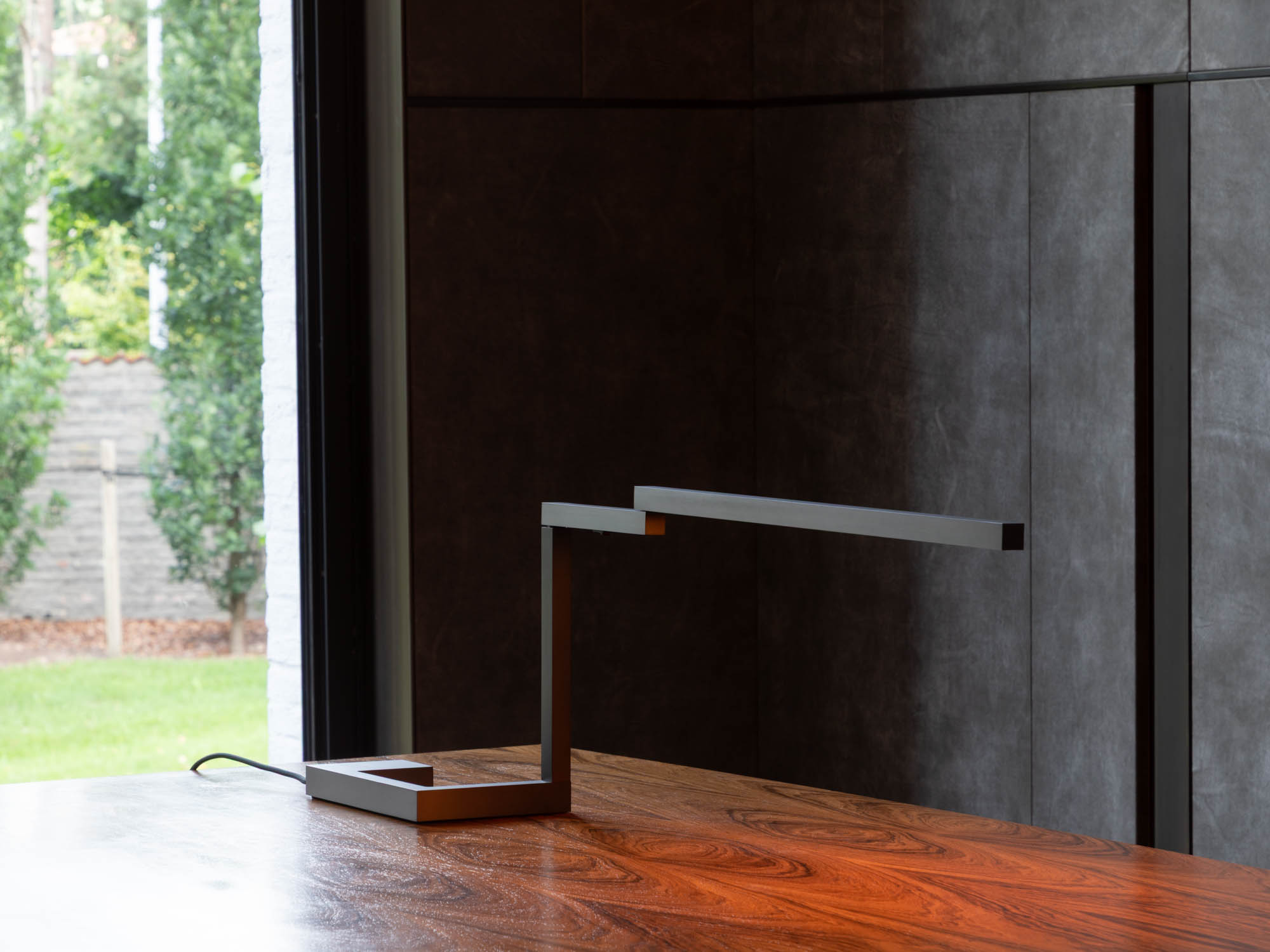 The Untitled table lamp, with its dark structure placed on a varnished wooden desk facing a black wall.