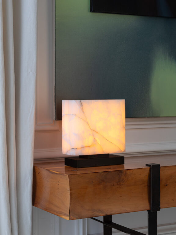 Auf einem Holzmöbel platziert, beleuchtet die Tischleuchte Embrun LP ein großes grünes Bild im Hintergrund.
