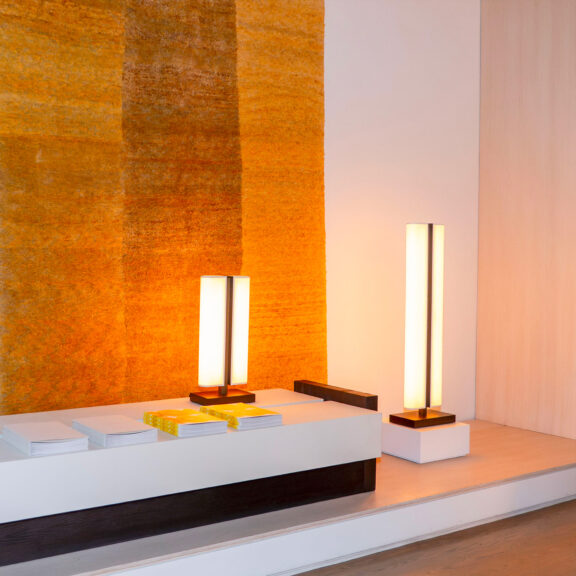 Illuminating an orange tapestry, a Tennessee LP table lamp and a Tennessee LH floor lamp—both designed by Glenn Sestig for Ozone—are placed on a white piece of furniture.